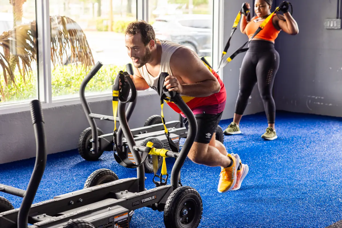 Man pushing a weighted sled while a woman trains with TRX straps at SWEAT440