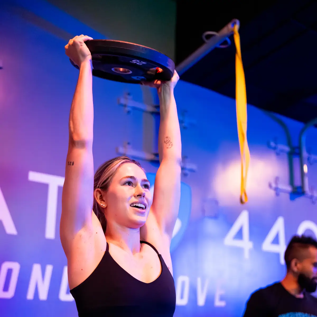 Woman pressing a weight plate overhead while a SWEAT440 coach looks on