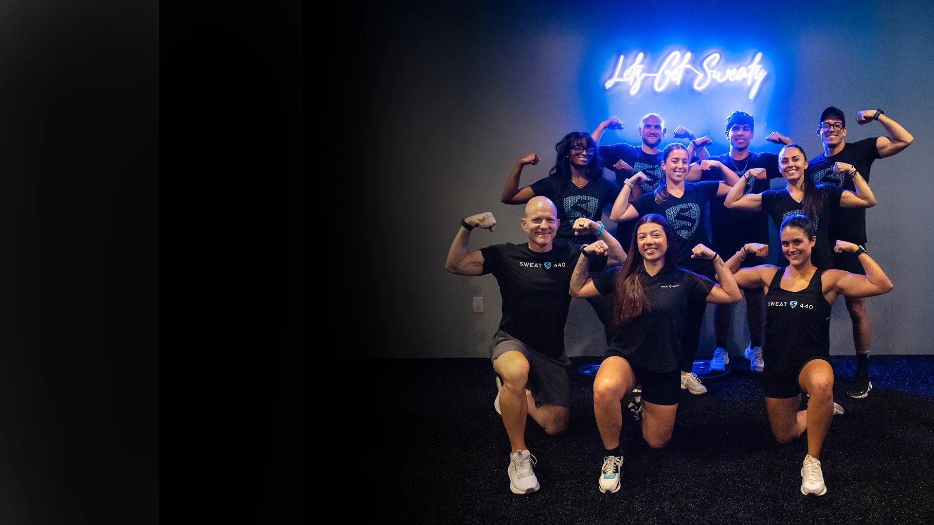 SWEAT440 coaches and members flexing together in front of the studio neon sign